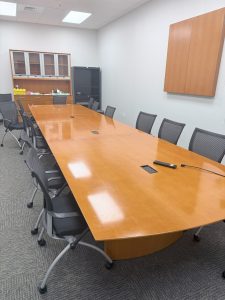 Large conference table surrounded by black office chairs in a meeting room with wooden cabinets and gray walls.