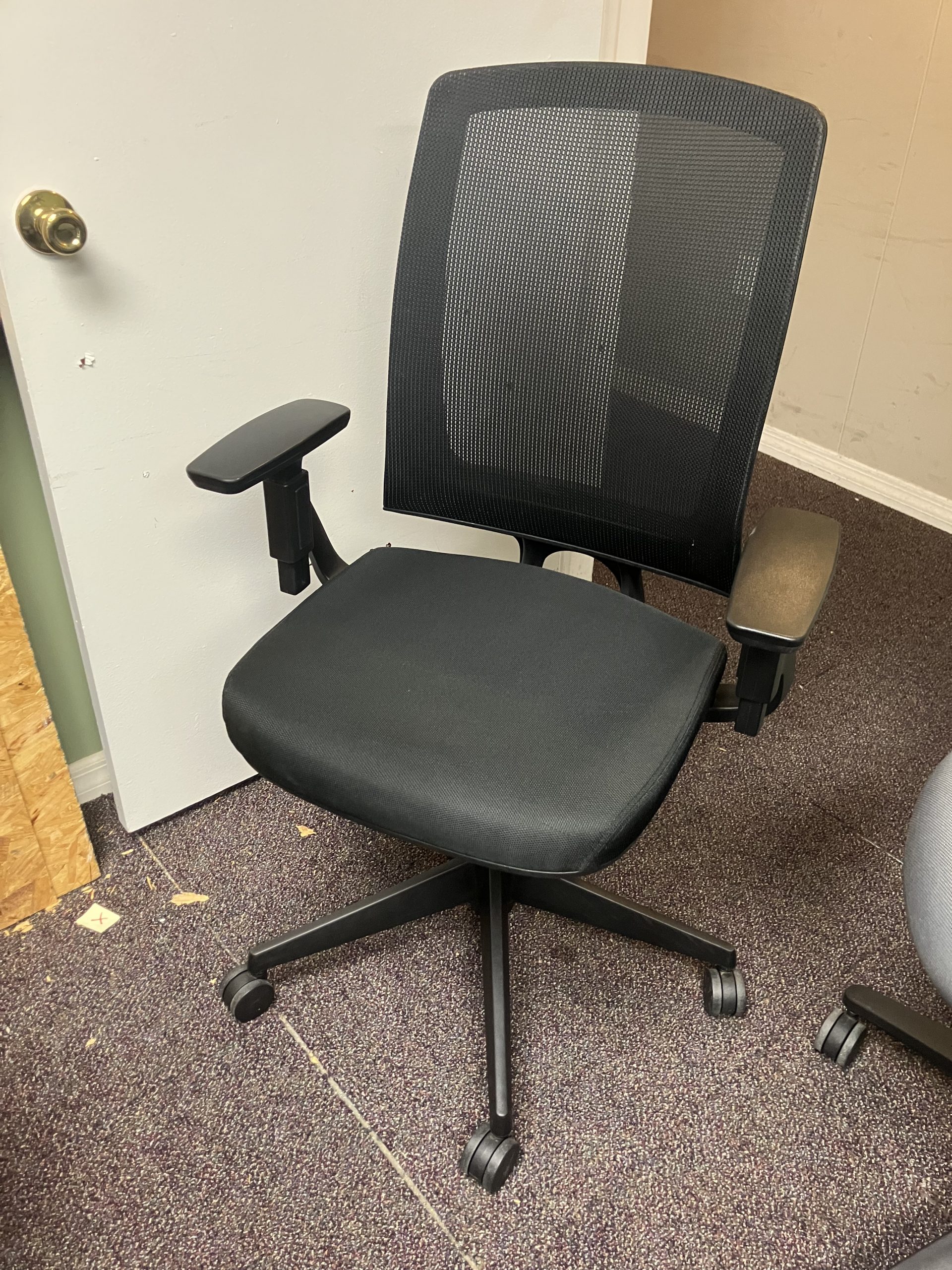 Ergonomic black office chair with a mesh back and padded seat on a five-wheel base in a carpeted room by a white door.